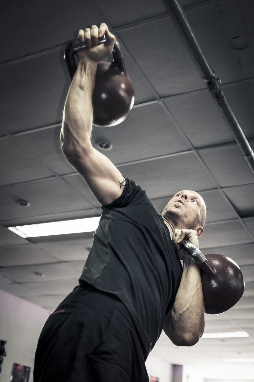 En person utför en press över huvudet med två kettlebells, vilket demonstrerar en övning som bygger styrka och stabilitet i axlarna.