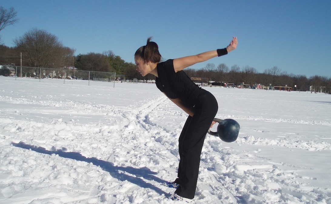 En person i svarta träningskläder utför en kettlebell-övning i snömiljö, vilket visar den direkta kopplingen mellan styrketräning och förberedelser för vintersport.